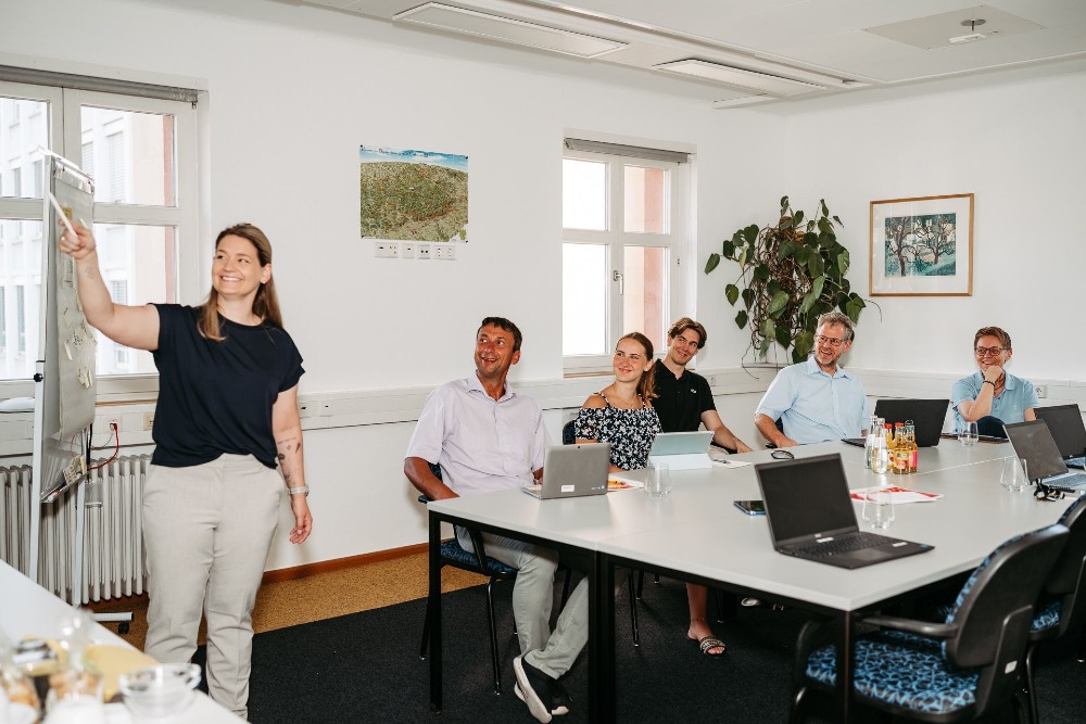 Eine Frau steht an einer Tafel und zeigt fünf Mitarbeitern, die an einer Tischrunde sitzen ihre gesammelten Notizen