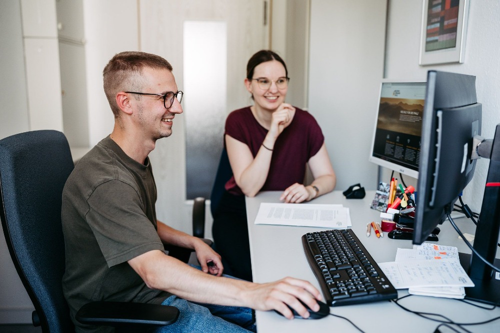 Zwei Personen sitzen am Schreibtisch. Dabei schauen beide auf den Bildschirm eines Computers